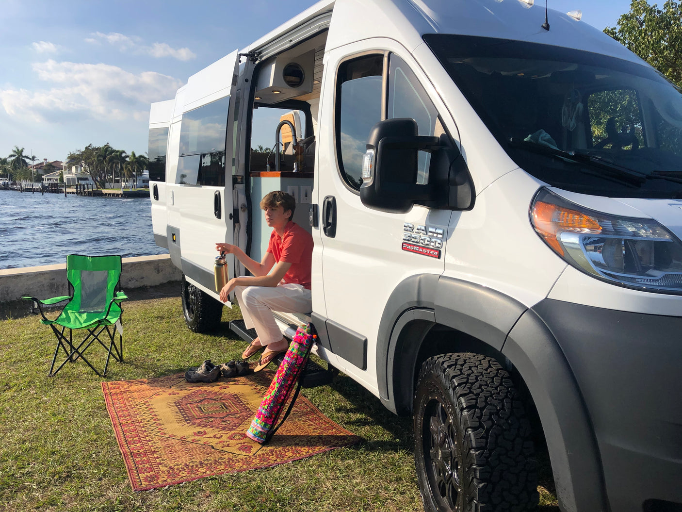 A young man relaxes in the side doorway of a white camper RV rental, parked on grass by the water, offering a serene van life retreat.