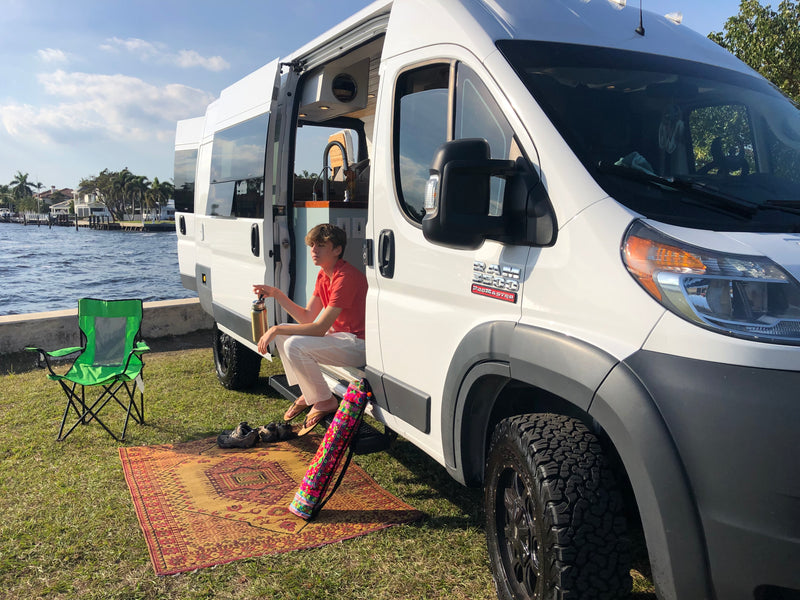 A young man relaxes in the side doorway of a white camper RV rental, parked on grass by the water, offering a serene van life retreat. — photo 20