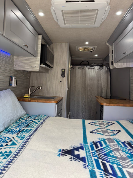 Compact RV rental interior: bed with blue patterned blanket, full kitchen with sink & microwave, and shower.