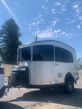 This Airstream Basecamp RV trailer, ideal for an rv rental adventure, shines under a sunny sky with trees and mountains.