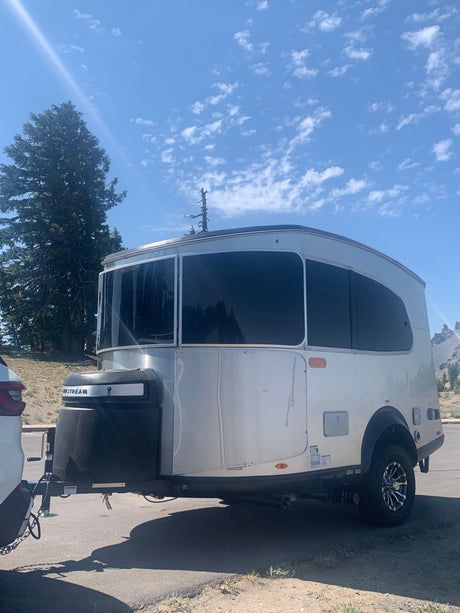 This Airstream Basecamp RV trailer, ideal for an rv rental adventure, shines under a sunny sky with trees and mountains.