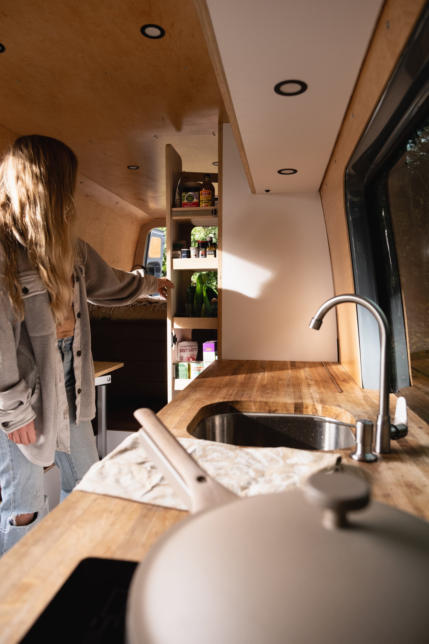 Cozy RV rental interior: A woman accesses the pantry in a beautifully designed camper van kitchen with wooden counters & sink.