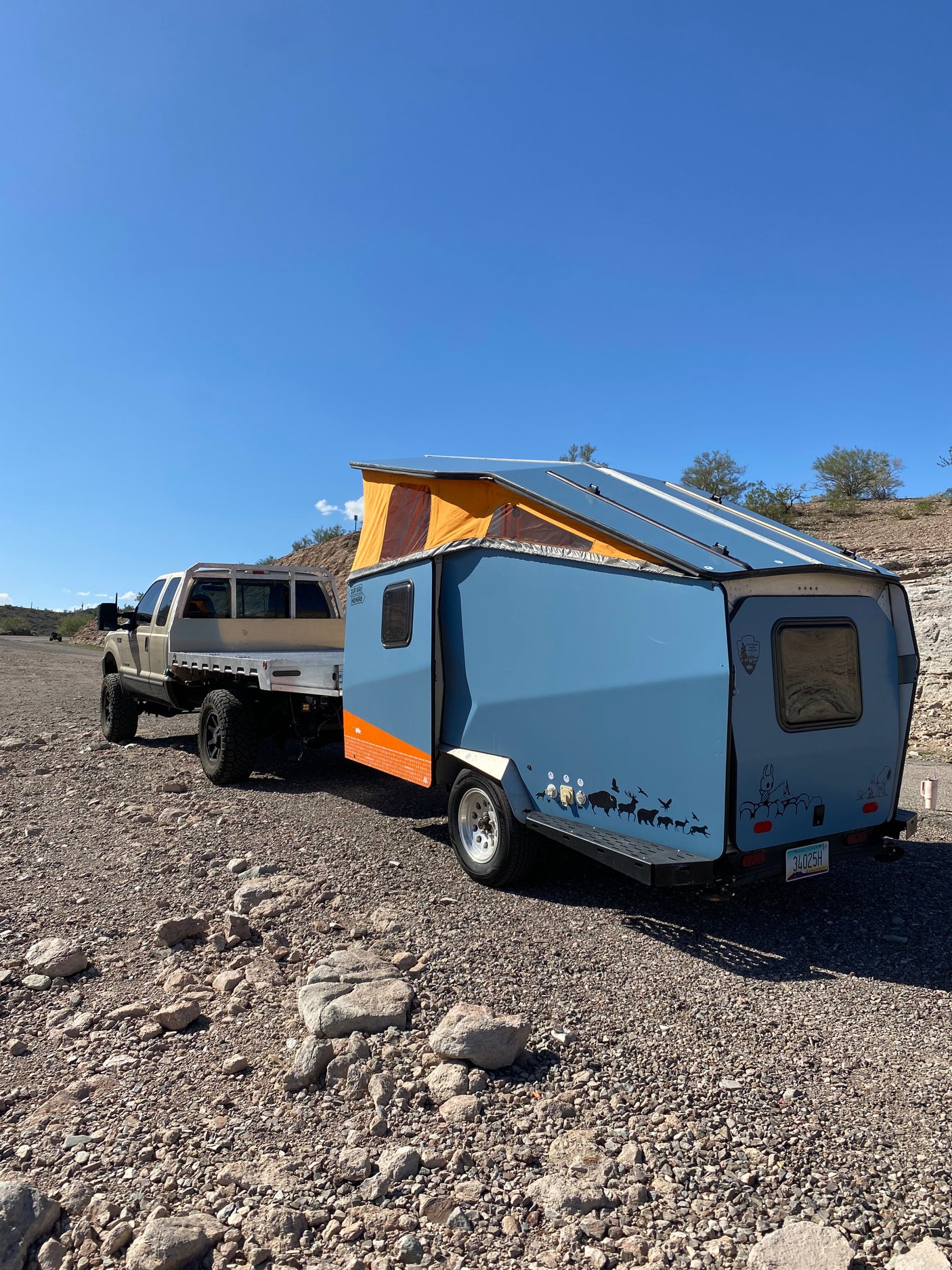 Adventure-ready RV rental: Truck towing a blue pop-top camper trailer on a rocky path under a clear blue sky, ideal for off-road travel.