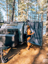 RV rental for camping: Woman by a modern teardrop trailer & ROAM storage box, using an outdoor shower tent by the lake.