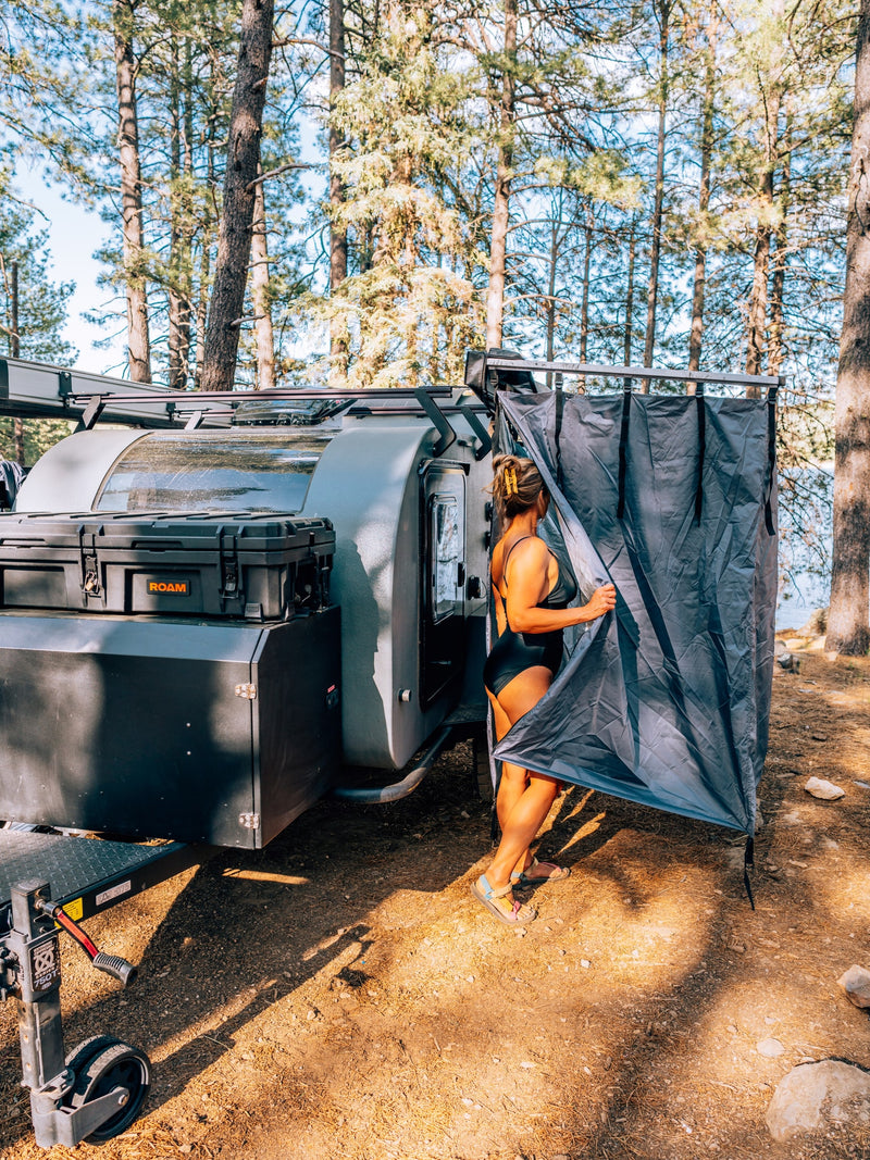 RV rental for camping: Woman by a modern teardrop trailer & ROAM storage box, using an outdoor shower tent by the lake. — photo 11
