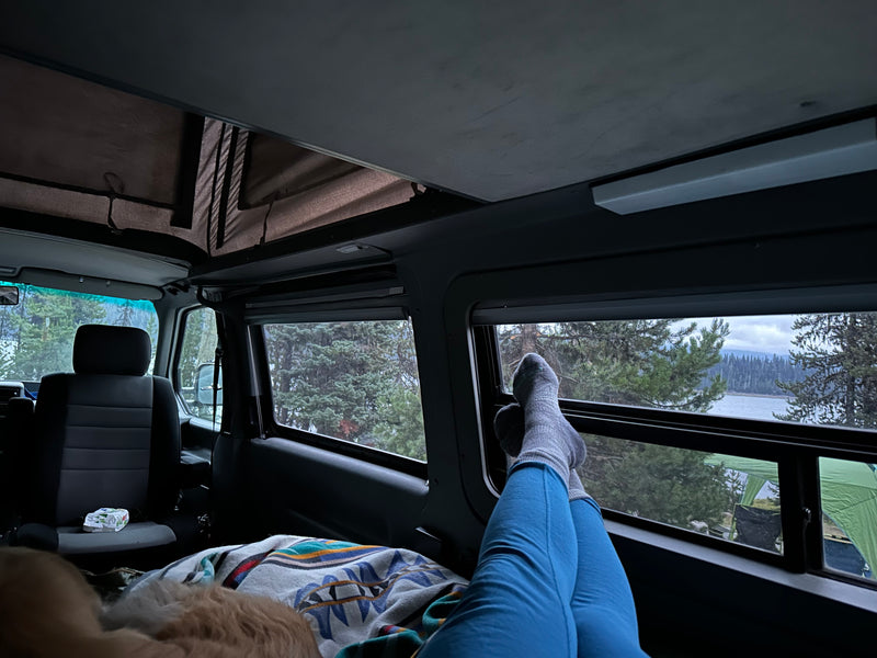 Cozy rv rental: person relaxing inside a camper van with feet up, enjoying a serene lake and forest view. — photo 11