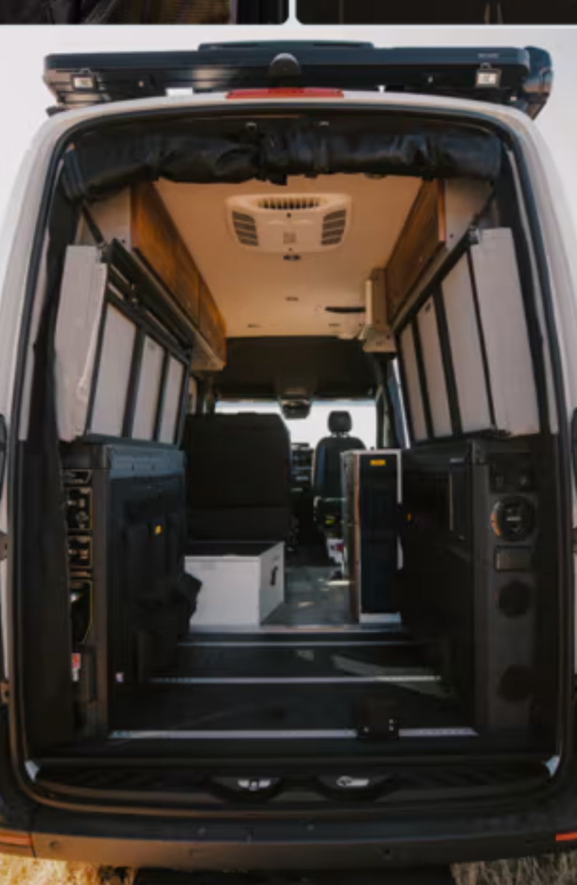 View inside an RV rental: a custom camper van interior with modular storage, seating, and an overhead AC unit from the rear doors.