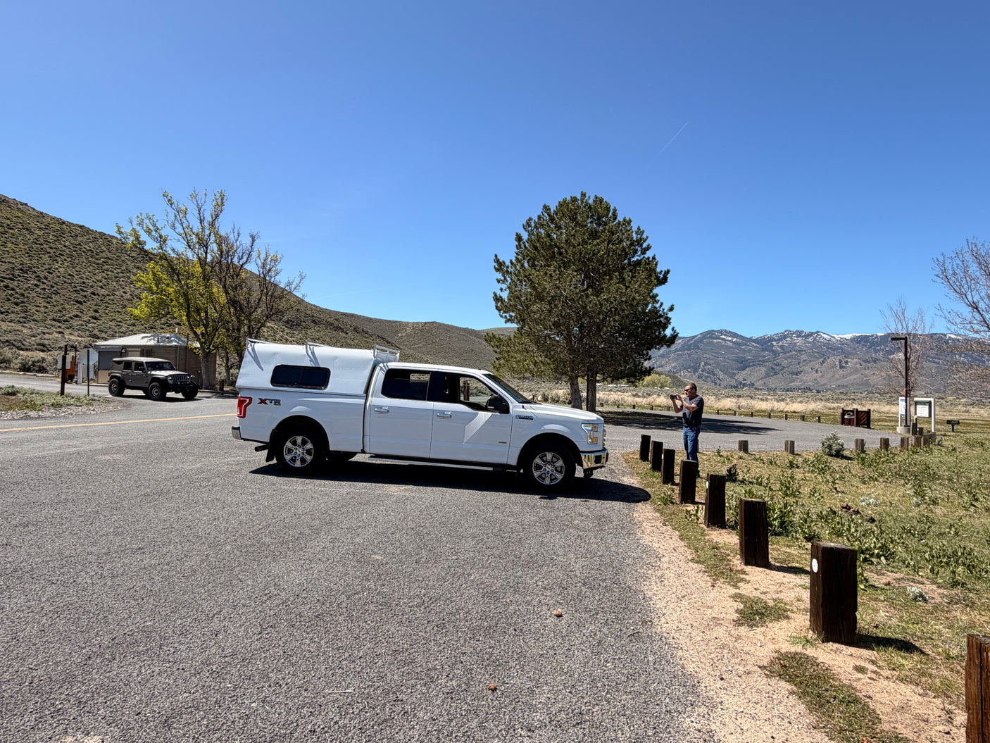 A white pickup truck with RV camper shell, an ideal RV rental for exploring scenic mountain roads. A man takes photos of snowy peaks.