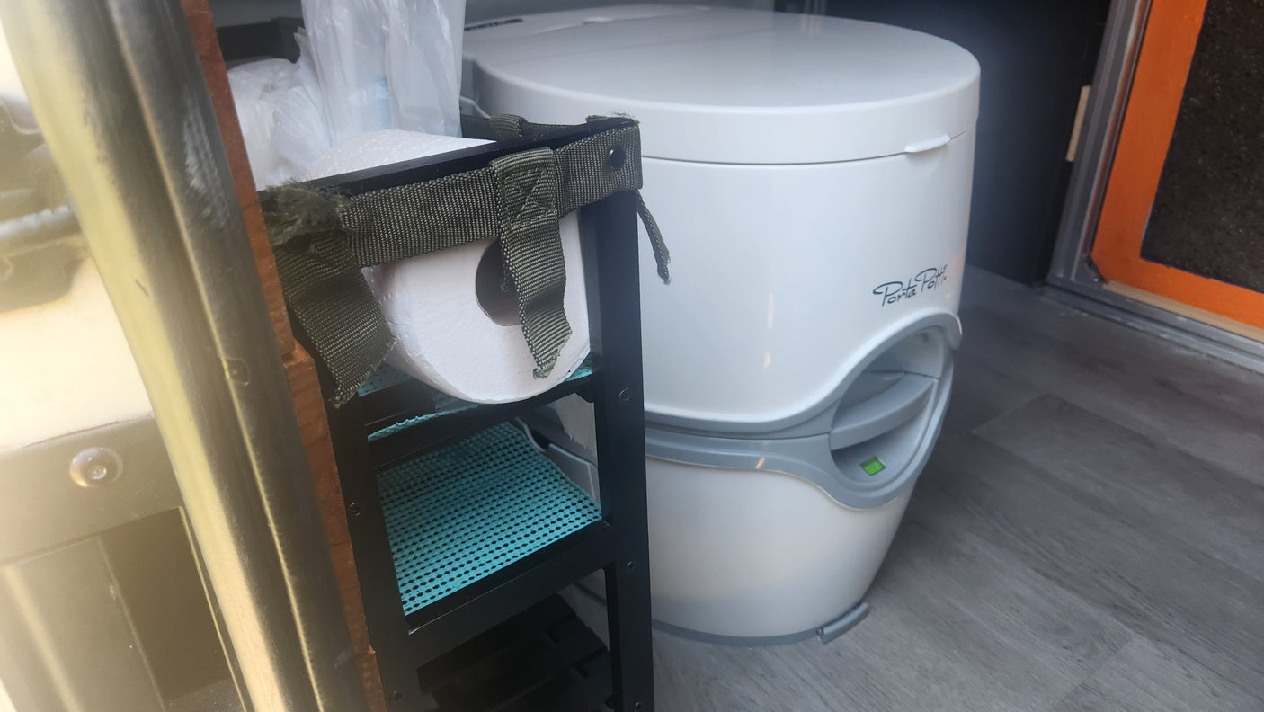 RV rental: White Porta Potti portable toilet beside a compact shelving unit with toilet paper.