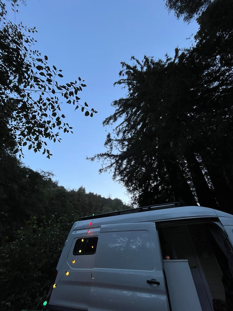 RV rental campervan at dusk, adorned with colorful string lights. A white van is parked among dark trees under a clear blue sky.