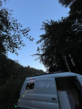 RV rental campervan at dusk, adorned with colorful string lights. A white van is parked among dark trees under a clear blue sky.