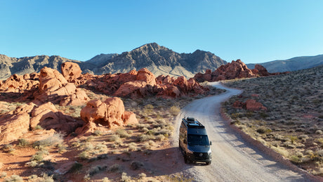 RV rental van on a desert road, surrounded by stunning red rock formations & mountains under a clear sky. Ideal for adventure travel.