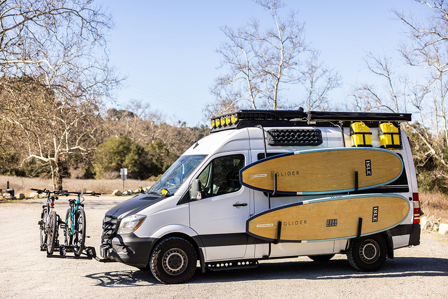 An adventure-equipped RV rental van, featuring bikes on the front, paddleboards on the side, and rooftop storage.