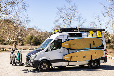 An adventure-equipped RV rental van, featuring bikes on the front, paddleboards on the side, and rooftop storage.