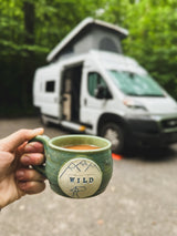 A hand holds a green "WILD" mug with coffee. An RV rental camper van is blurred in the forest background. Ideal for nature trips.