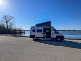 Scenic RV rental: a silver camper van with pop-top open, parked on asphalt beside a lake under a clear blue sky.