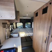 Interior of an RV rental with a clean kitchen counter & sink on the left, leading to a comfortable bed in the rear. Storage on right.