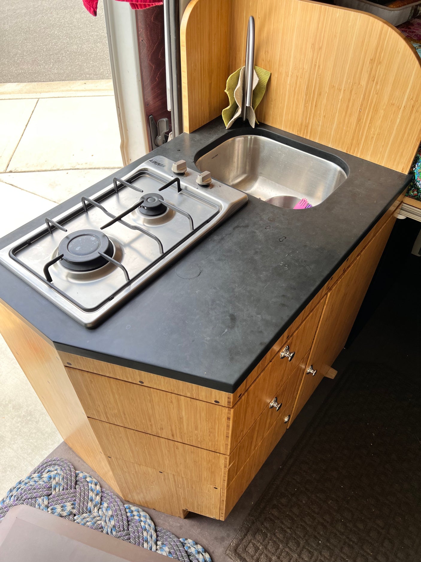 This RV rental features a compact kitchen with a two-burner stove, sink, black countertop, and bamboo cabinetry.