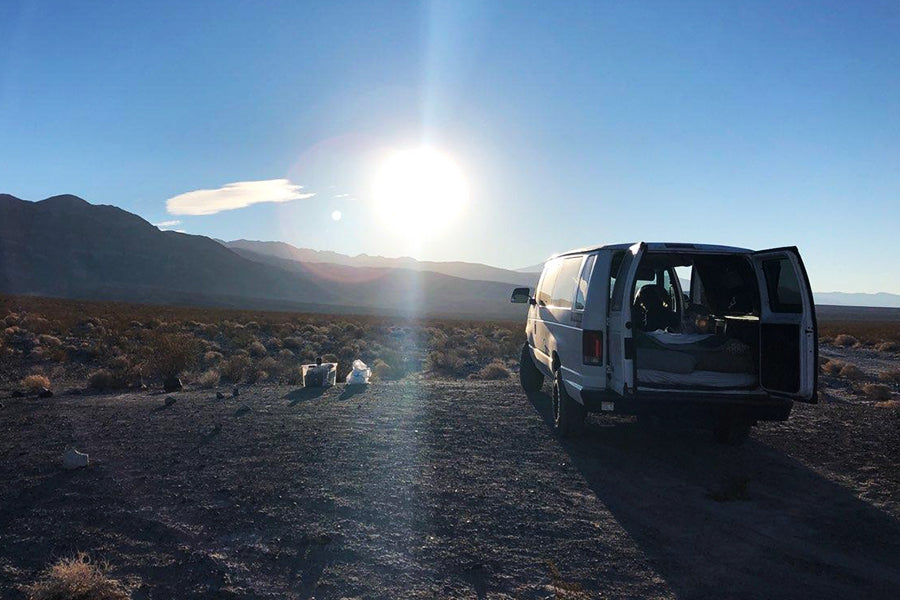 White RV rental van with open doors parked on a dirt road in a sunny desert landscape at golden hour, mountains afar. Camping setup visible.