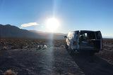 White RV rental van with open doors parked on a dirt road in a sunny desert landscape at golden hour, mountains afar. Camping setup visible.