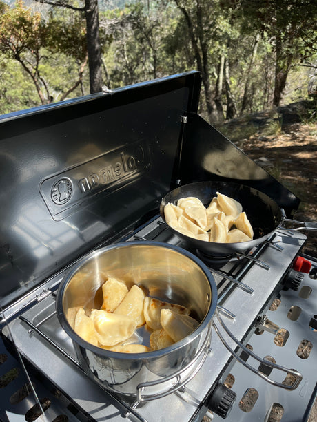 A portable camping stove holds two pans of pierogi in a natural outdoor setting, ideal for an RV rental trip.