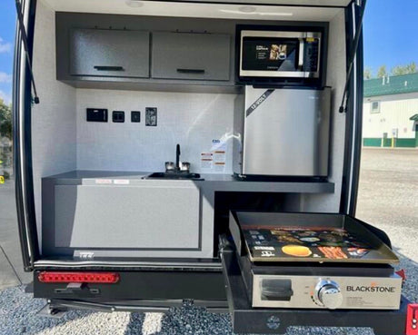 Compact RV rental outdoor kitchen featuring a microwave, 12V fridge, sink, and pull-out Blackstone griddle.