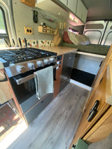 This RV rental features a compact kitchen with a modern stove, oven, wood countertop, fridge, and a cozy bed in the background.