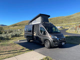 This RV rental, a grey camper van with its pop-top extended and door open, is parked at a scenic campsite.