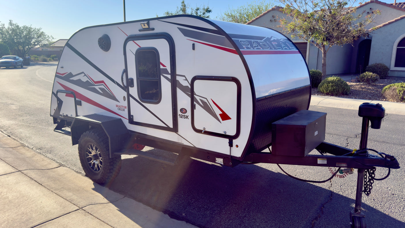 White Braxton Creek 12SK teardrop rv rental trailer, featuring off-road tires and black/red graphics, parked on pavement.