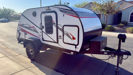 White Braxton Creek 12SK teardrop rv rental trailer, featuring off-road tires and black/red graphics, parked on pavement.