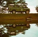 An iconic green Land Rover, ideal for an RV rental adventure, perfectly mirrored in calm water, ready for exploration.