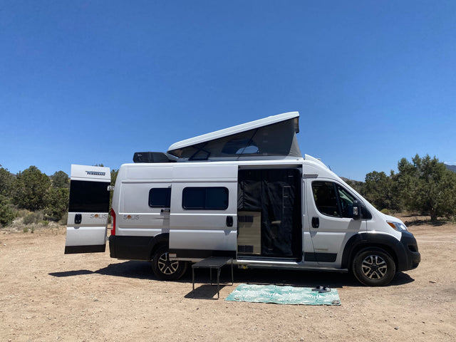 White Winnebago Solis RV rental with pop-top open, set up for camping in nature. Mat and table outside.