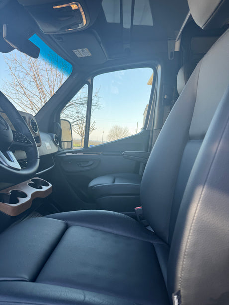 Inside a modern rv rental van. View of front seats, steering wheel, custom cup holder, and sunlit outdoor scenery.