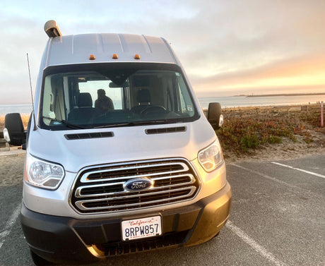 A silver Ford Transit RV rental is parked at a California beach lot during golden hour, ideal for travel.