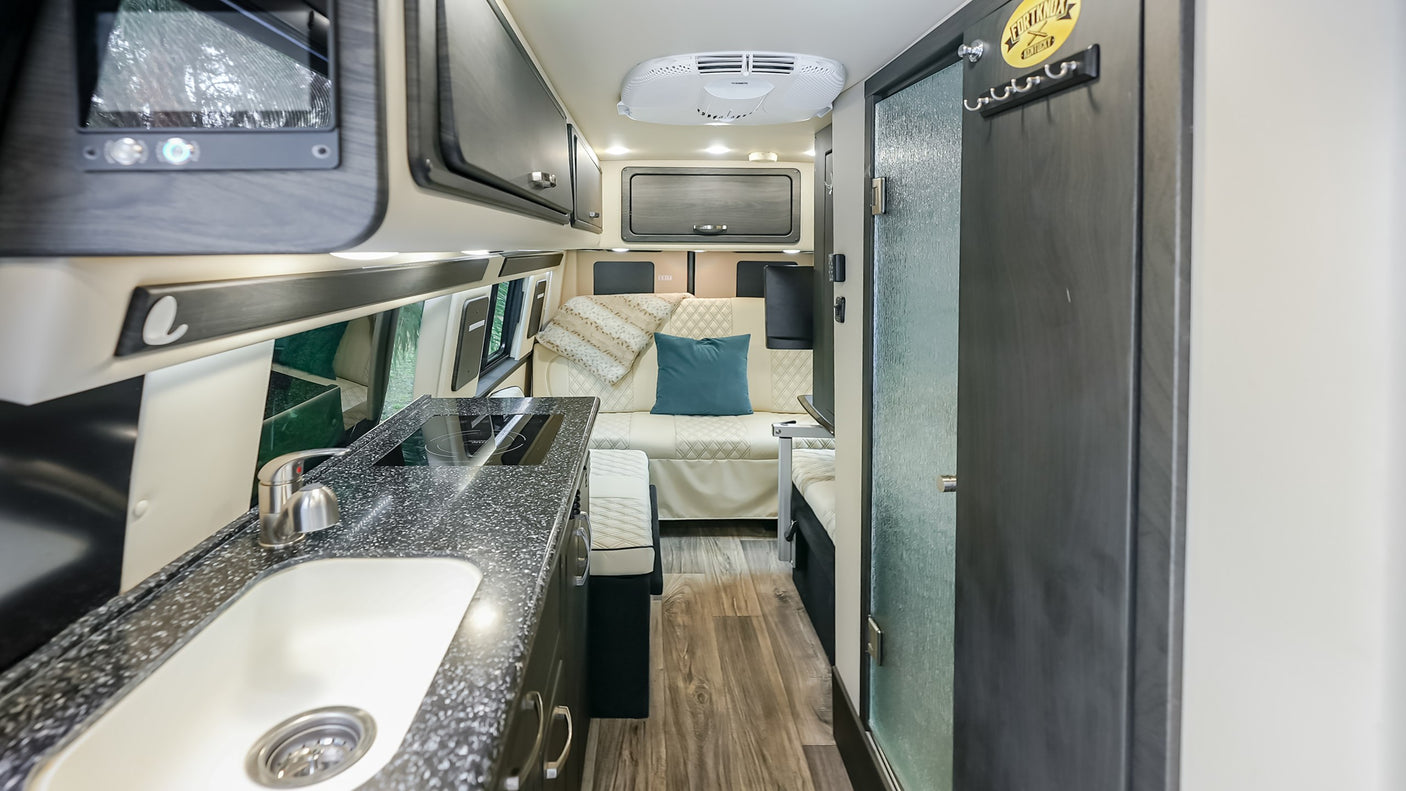 A sleek rv rental interior shows a kitchen with a sink, cooktop, lounge seating with pillows, overhead cabinets, and ceiling AC.