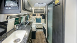 A sleek rv rental interior shows a kitchen with a sink, cooktop, lounge seating with pillows, overhead cabinets, and ceiling AC.