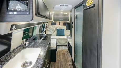 A sleek rv rental interior shows a kitchen with a sink, cooktop, lounge seating with pillows, overhead cabinets, and ceiling AC.