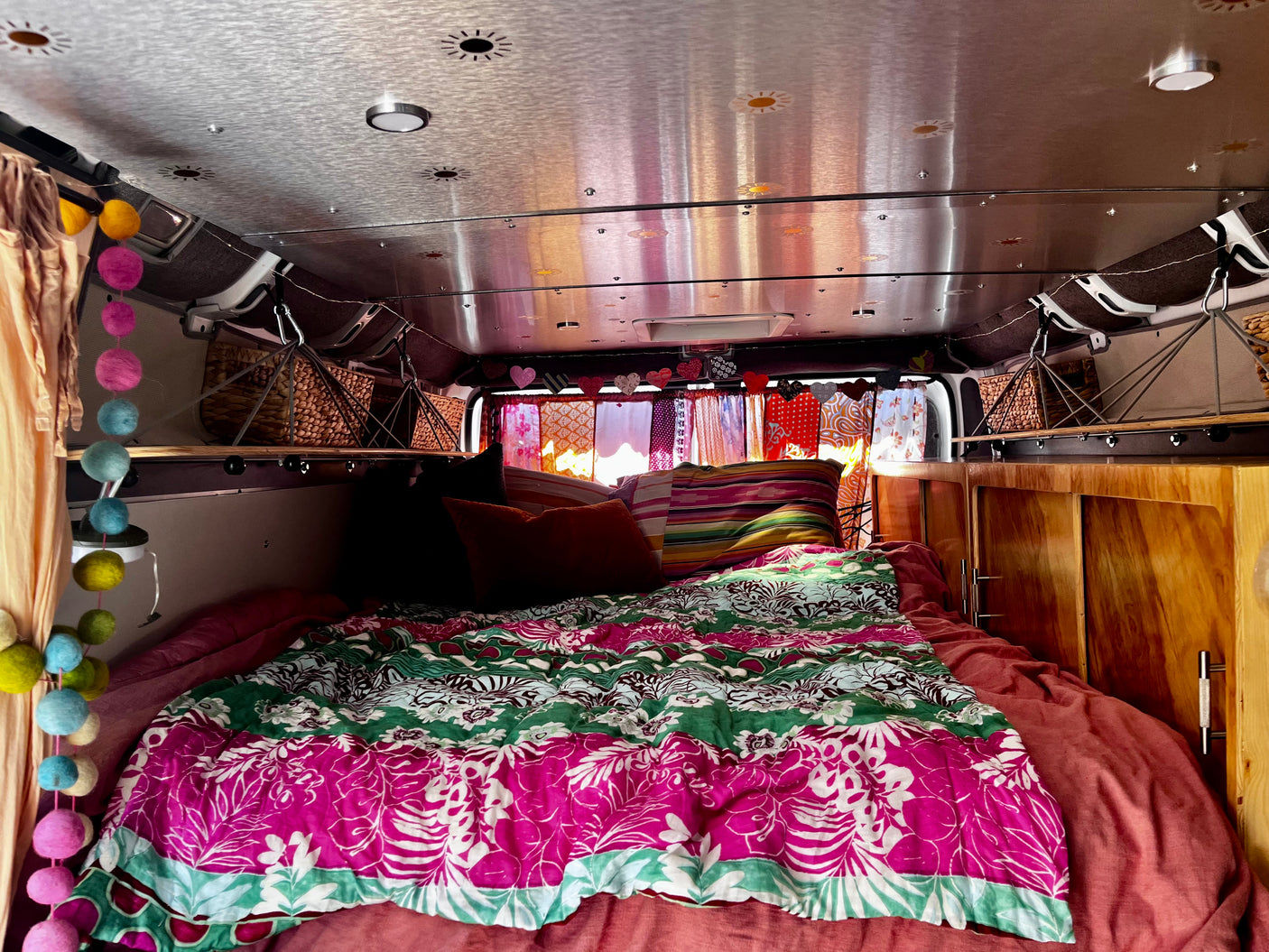 Inside a vibrant RV rental: cozy bed with pink/green quilt, wooden cabinets, and string lights create a warm, inviting space.