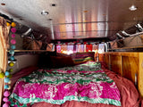 Inside a vibrant RV rental: cozy bed with pink/green quilt, wooden cabinets, and string lights create a warm, inviting space.