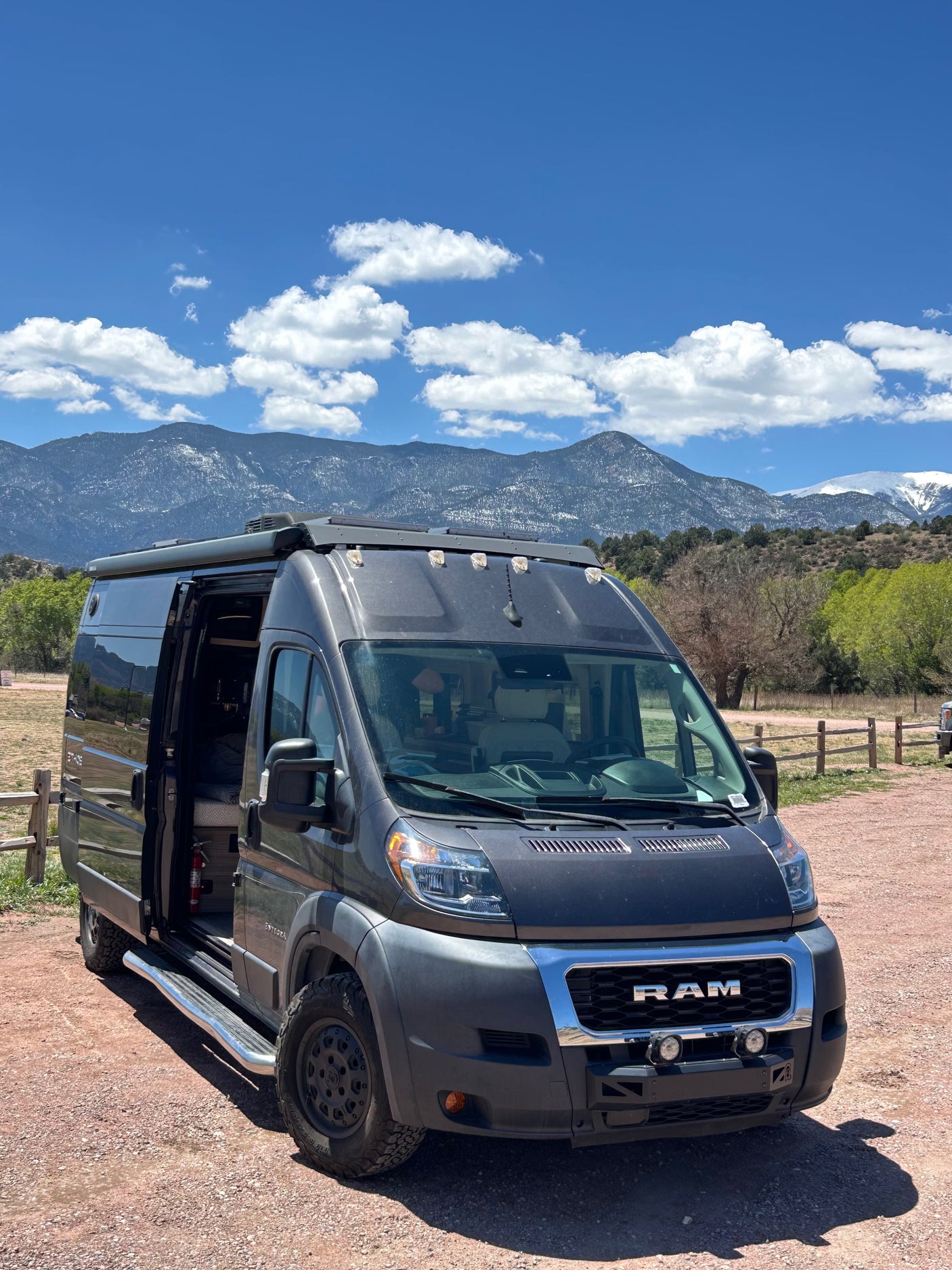 Adventure-ready dark grey RAM ProMaster RV rental van with off-road tires, awning, and snowy mountains under a blue sky.