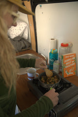 A person stirs food in a pan on a portable stove inside a compact RV rental kitchen, with a book and bottles nearby.