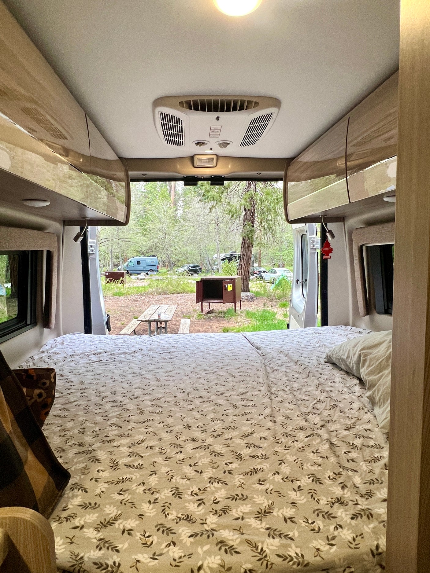 Inside a comfy RV rental, the bed is made, and open doors reveal a peaceful campsite with trees, a picnic table, and other vehicles.