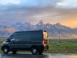An RV rental camper van faces majestic Grand Teton mountains at sunset. Snow-capped peaks glow under a dramatic sky.