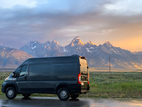 An RV rental camper van faces majestic Grand Teton mountains at sunset. Snow-capped peaks glow under a dramatic sky.