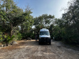 A white camper van, possibly an RV rental, parked at a rustic campsite with a picnic table and a person standing nearby, surrounded by trees.