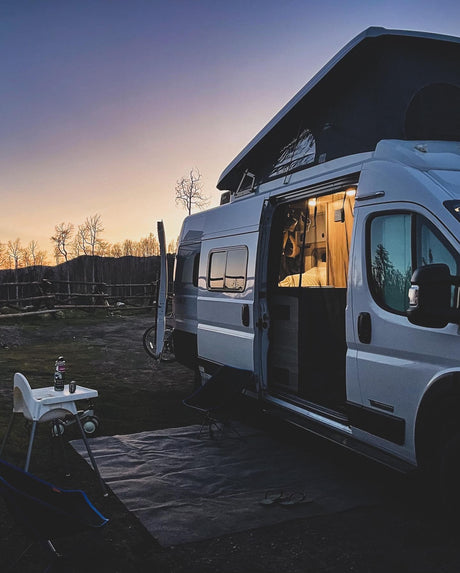 Perfect RV rental camping scene at dusk: white camper van with pop-top open, illuminated interior, and outdoor family setup.