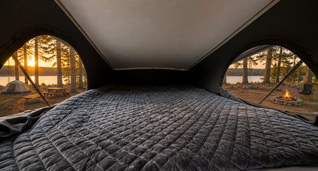 Cozy bed in an RV rental looking out at a beautiful lakeside campsite with a sunset, tent, and campfire. Ideal for glamping.