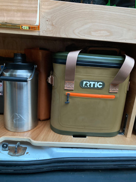 RV rental setup: olive RTIC cooler & silver mountain-logo water bottle on custom wood storage shelves.