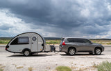 A gray SUV tows a sleek teardrop RV rental camper, parked on gravel under dramatic storm clouds. Ideal for outdoor adventures.