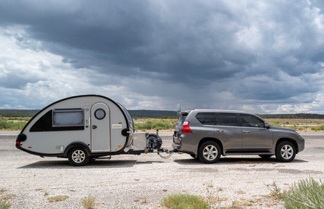 A gray SUV tows a sleek teardrop RV rental camper, parked on gravel under dramatic storm clouds. Ideal for outdoor adventures.